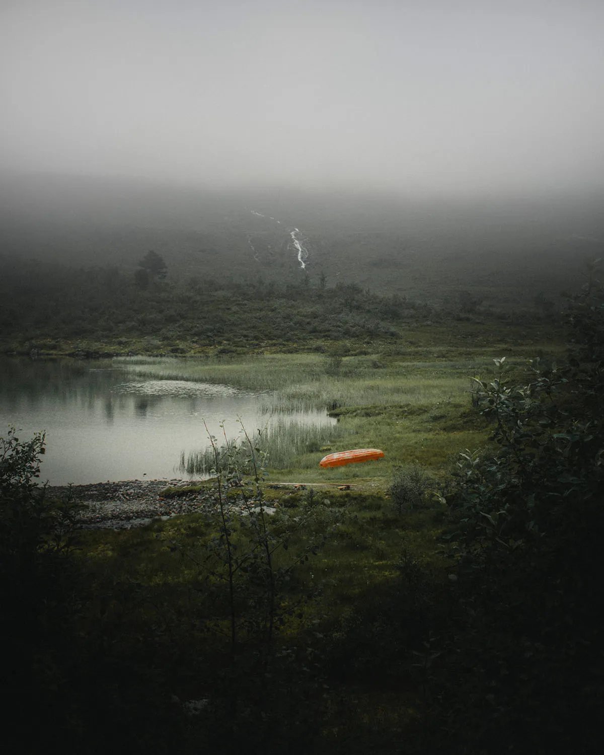 Foggy landscape with a still lake, grassy shore, and an upside-down orange canoe near the water.