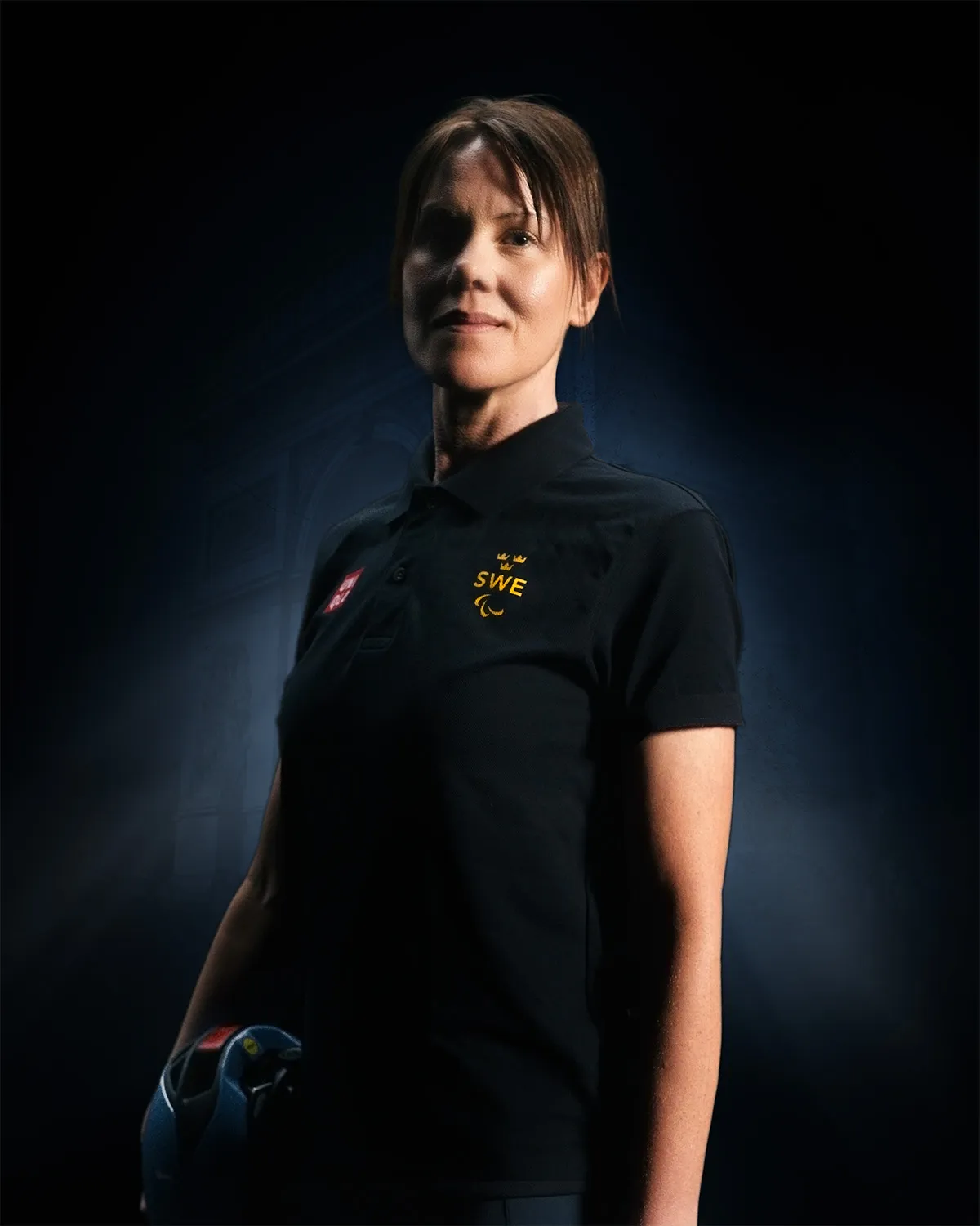 Confident woman wearing a black SWE polo shirt holding a blue helmet against a dark background.