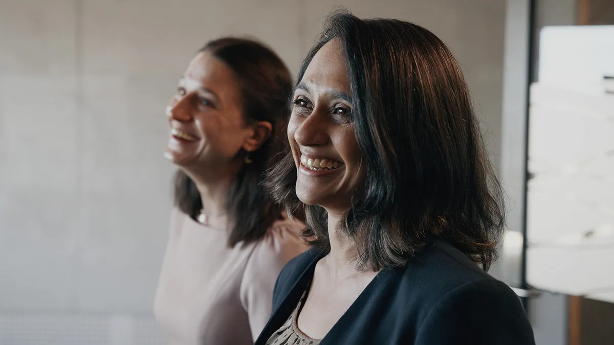 Two women smiling and looking towards the right in a softly lit indoor setting.