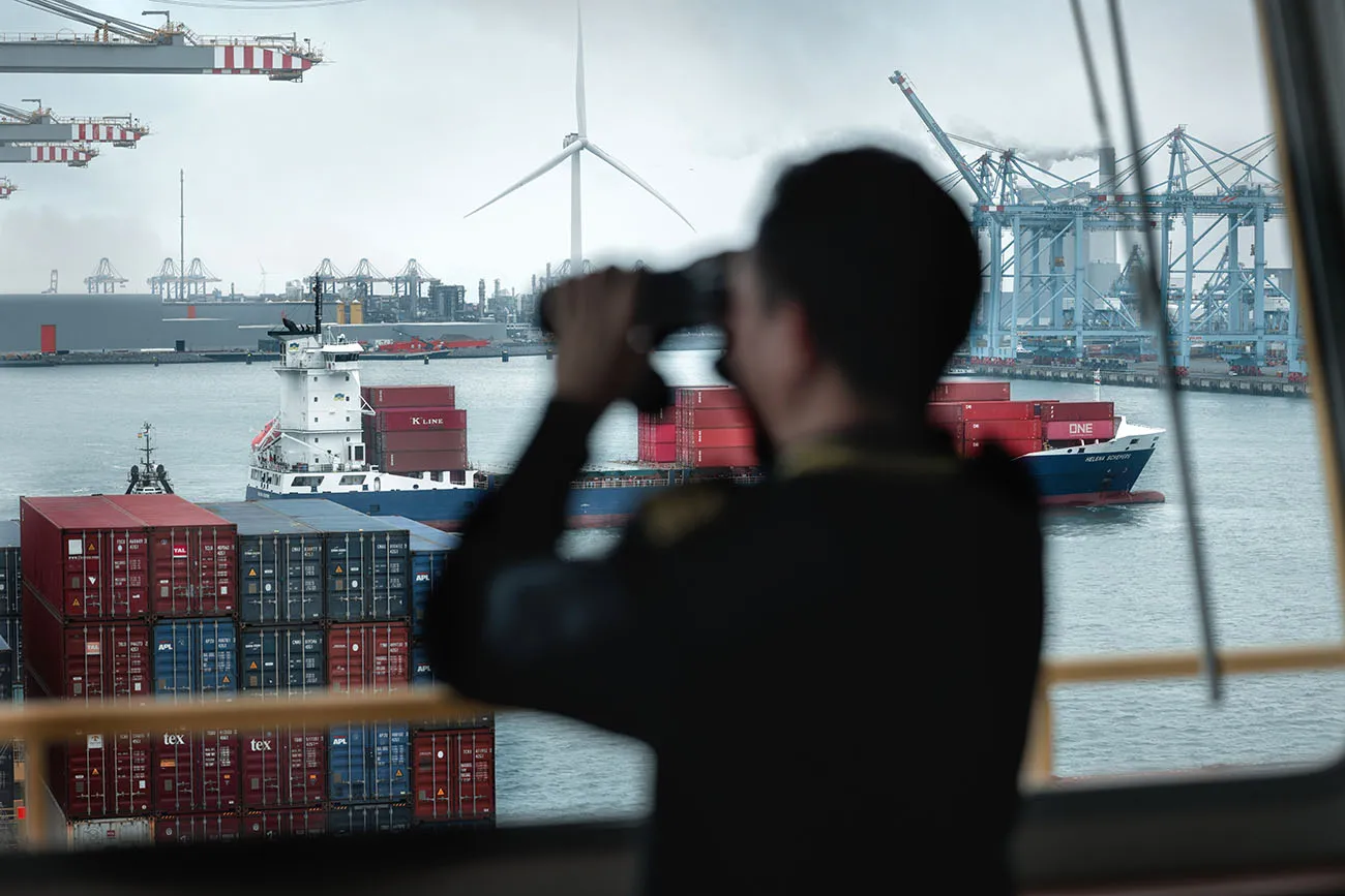 Silhouette of a person looking through binoculars at a busy port with container ships, cranes, and wind turbines in the background.