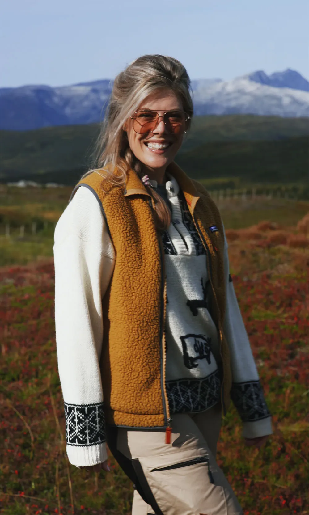 Smiling woman wearing orange-tinted sunglasses, a mustard fleece vest, and a patterned white sweater outdoors with mountains in the background.