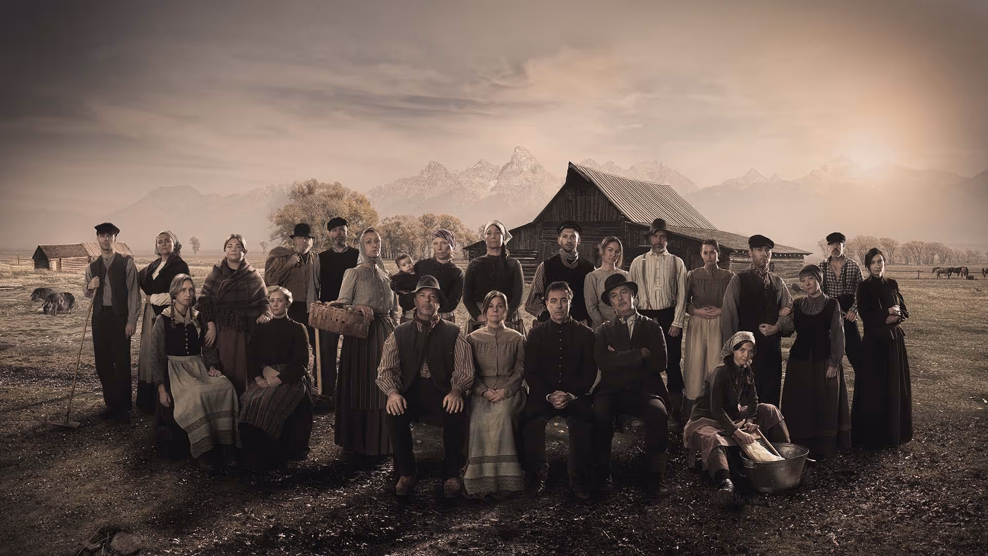 Black and white photo of a large group of pioneer settlers posing in front of a barn with mountains in the background during sunset.