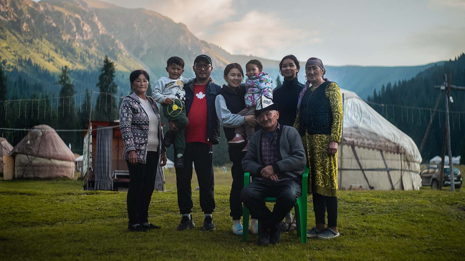 A multi-generational Kyrgyz family posing outdoors with traditional yurts and mountains in the background.