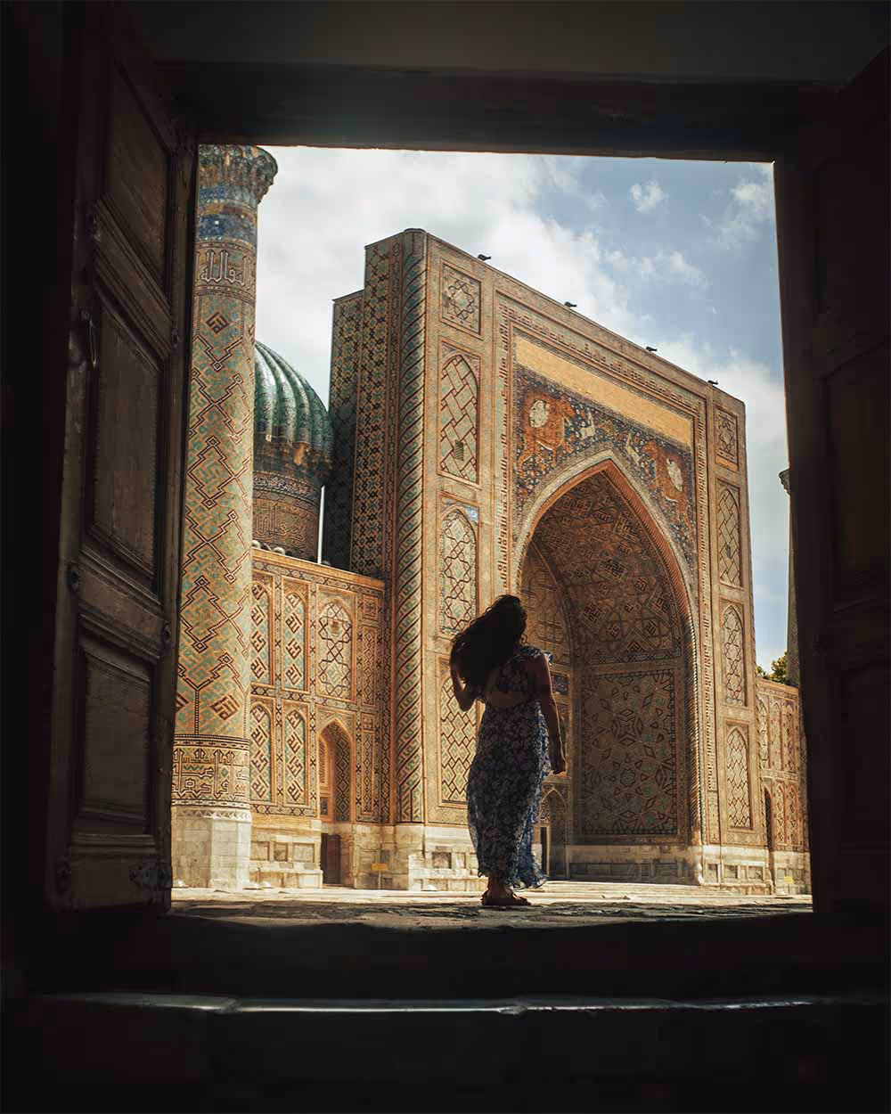 Silhouette of a woman in a floral dress walking toward an intricately tiled historic building with a large arch and green dome under a partly cloudy sky.