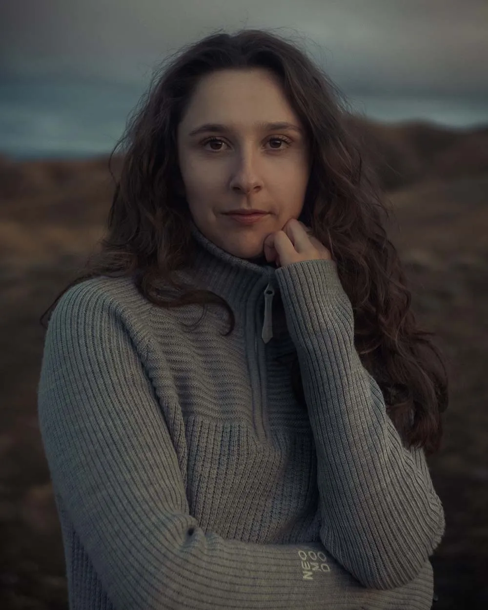 Woman with long dark curly hair wearing a gray knitted sweater outdoors with a mountainous background at dusk.