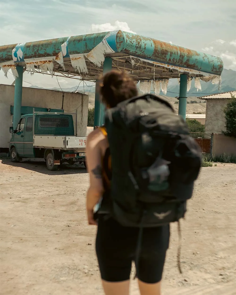 Backpacker with a large backpack standing in front of a rusty, dilapidated gas station canopy and an old truck.