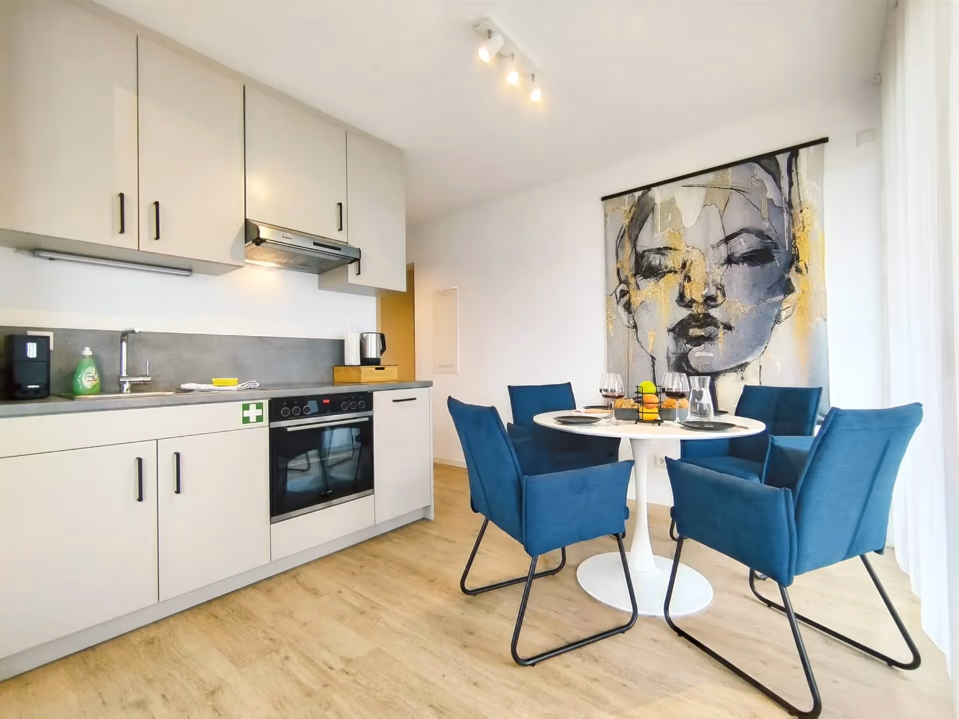 Moderne Apartmentküche mit Essbereich, runden Tisch, vier blauen Stühlen und großformatigem Wandbild im Hintergrund