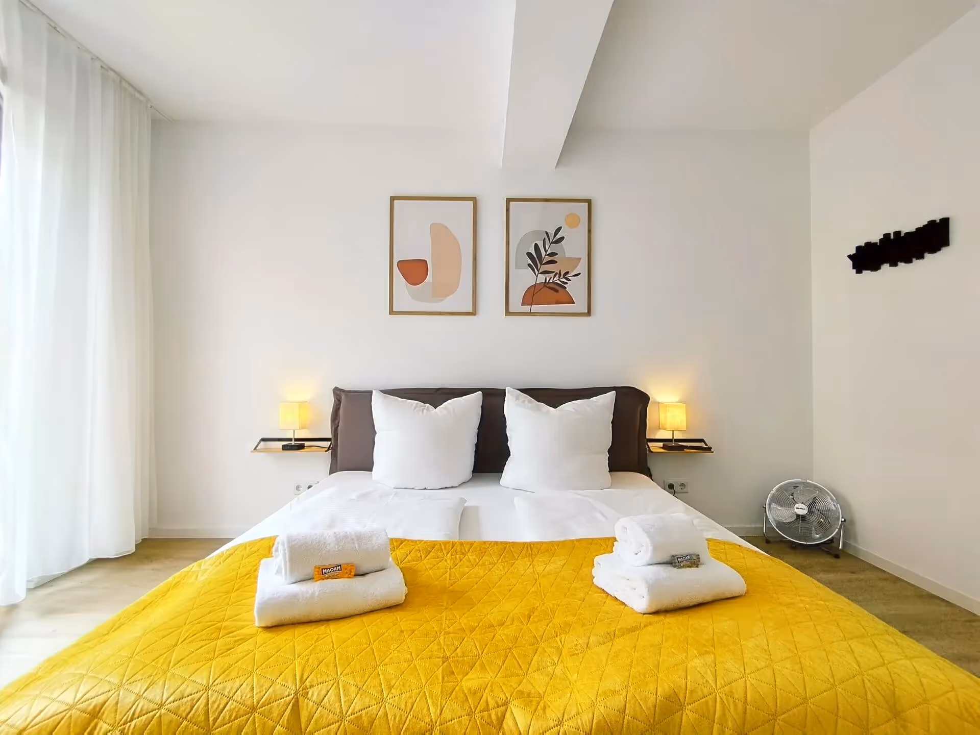 Schlafzimmer mit Doppelbett, gelber Tagesdecke, weißen Handtüchern und stilvollen Wandbildern – gemütlich und modern eingerichtet