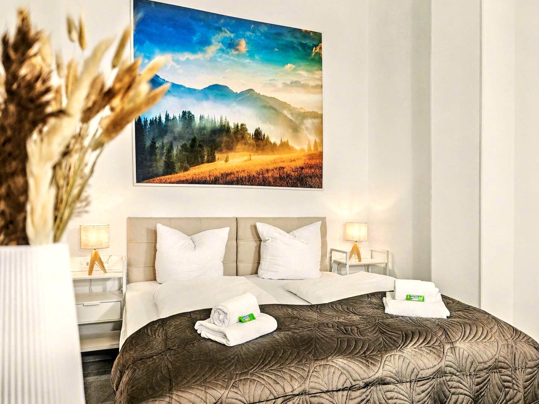 Auf dem Bild ist ein stilvoll eingerichtetes Schlafzimmer zu sehen. Das Doppelbett ist mit weißen Kissen und Decken sowie einer gesteppten, dunkelbraunen Tagesdecke bezogen. Zwei saubere Handtuchsets mit Seife liegen dekorativ auf dem Bett. Links im Vordergrund steht eine Vase mit Trockenblumen, im Hintergrund hängt ein großes Landschaftsbild mit Wald und Bergen im Sonnenlicht – es verleiht dem Raum eine warme, einladende Atmosphäre. Die Nachttische mit Lampen runden den gemütlichen Look ab.