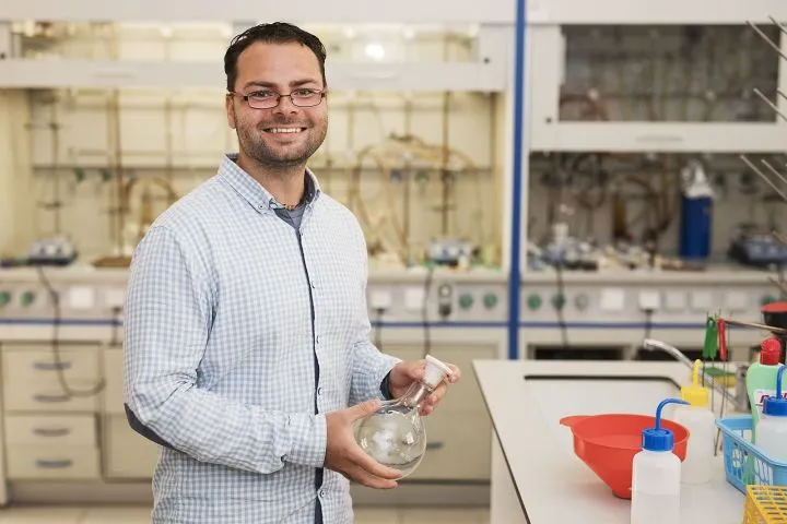 Man in glasses and checkered shirt holding glass flask in a chemistry lab with equipment in background.