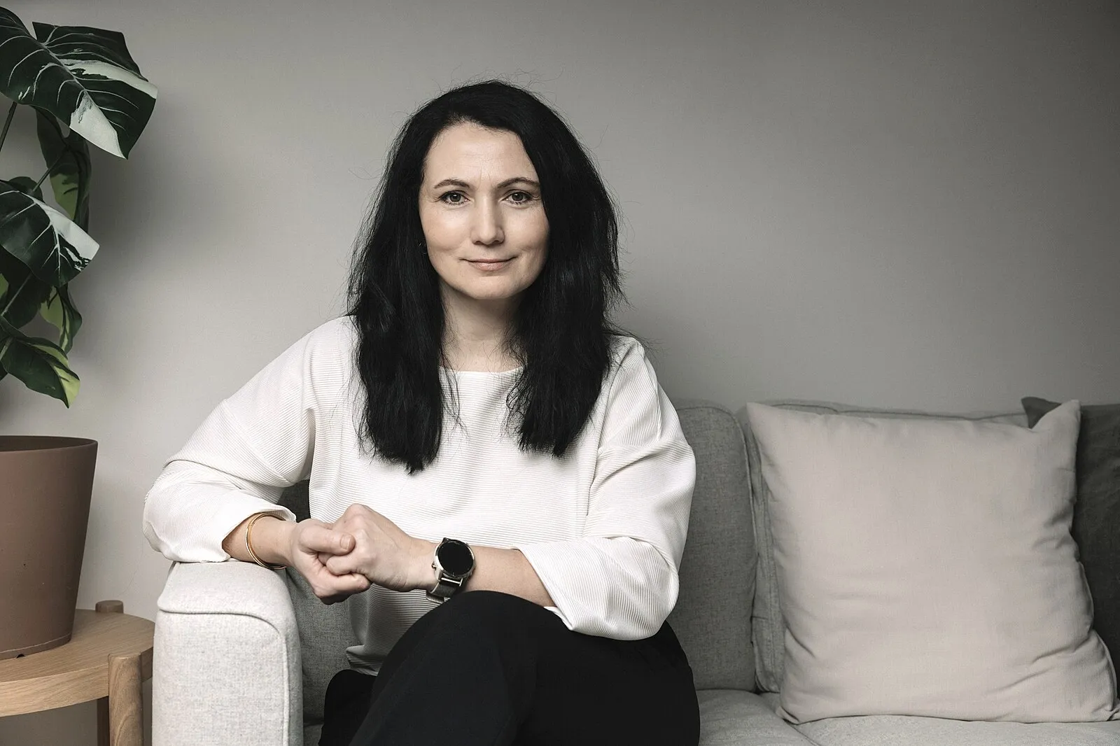 Woman with long dark hair wearing a white blouse sitting on a gray couch next to a plant.