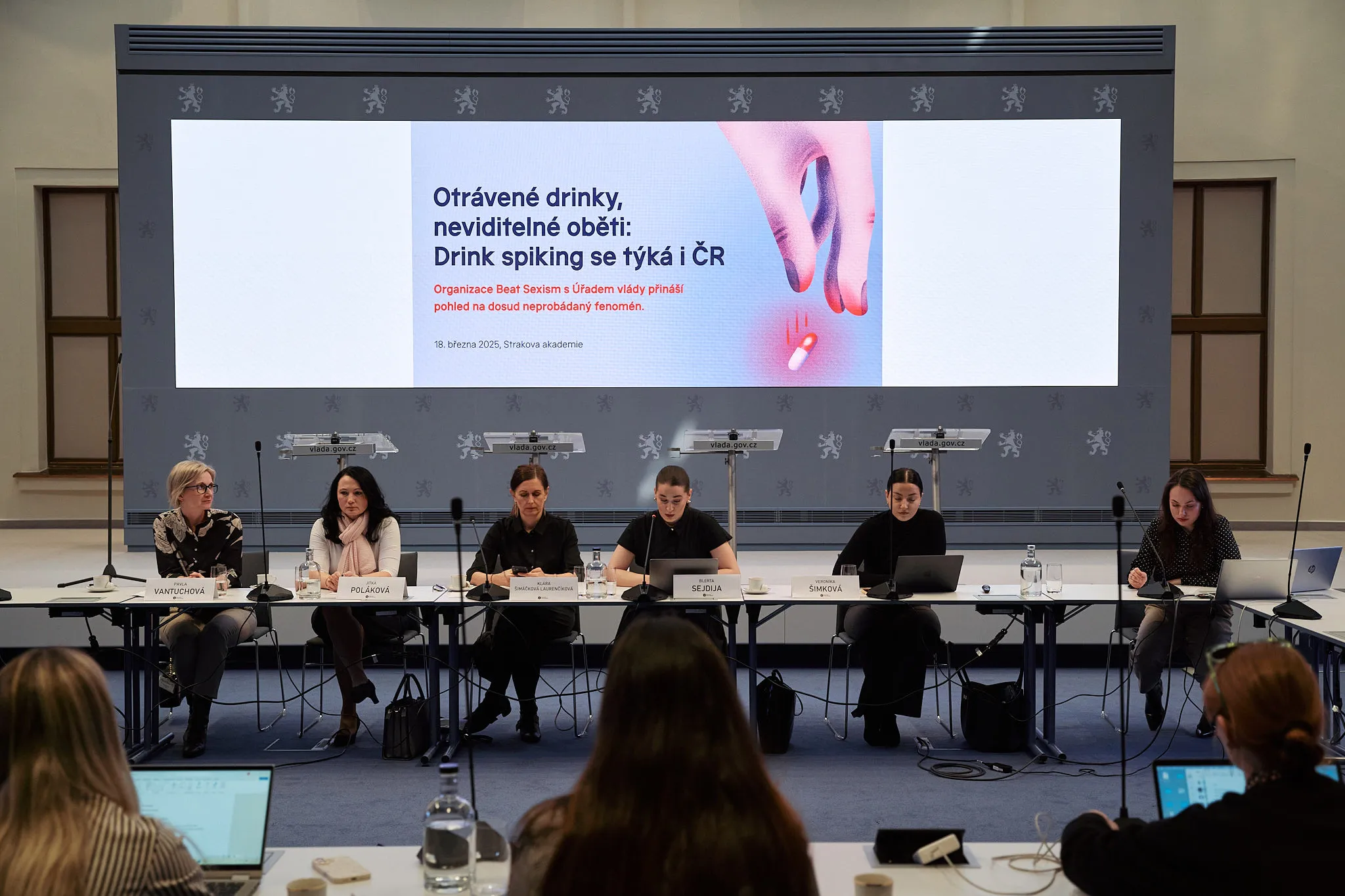 Panel of six women seated at a conference table with a screen about drink spiking behind them.