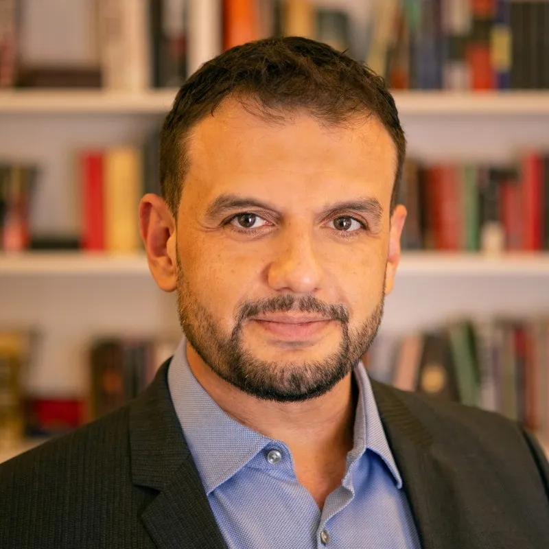Man with short dark hair and beard in blue shirt and dark suit jacket, blurred bookshelves behind him.