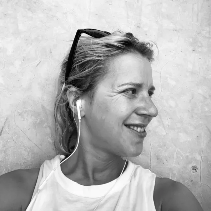 Smiling woman wearing earphones and sunglasses on her head against a textured wall.