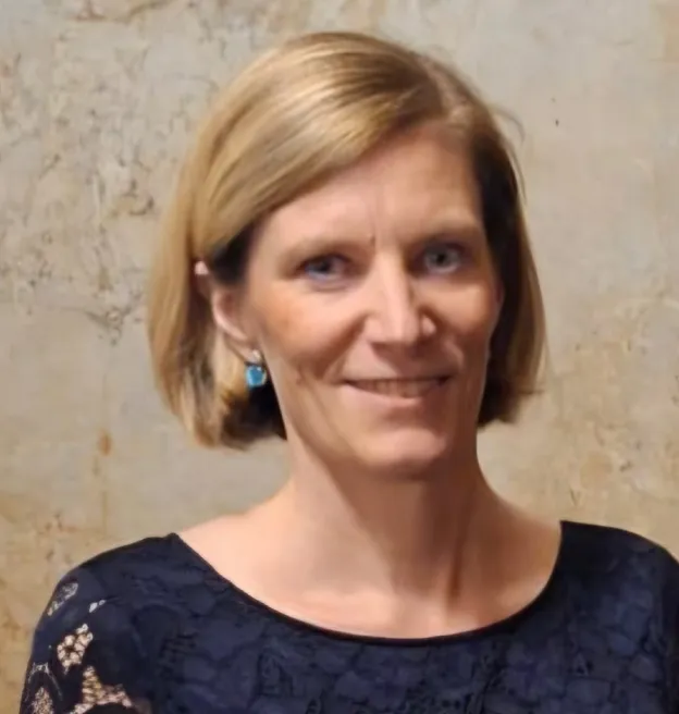 Smiling woman with short blonde hair wearing blue lace top and turquoise earrings against a neutral background.