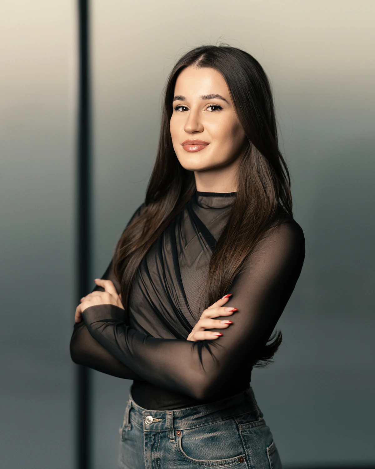 Young woman with long dark hair wearing a sheer black top and jeans, arms crossed, smiling slightly.