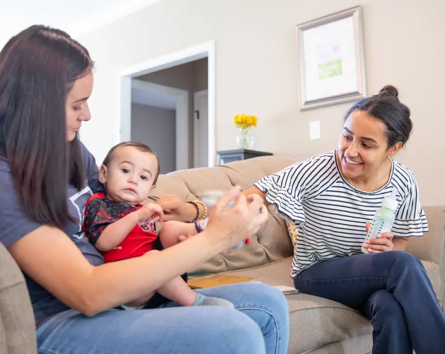 Two women sit on a couch, engaging with a baby between them, while one holds a water bottle and the other a drink. A flower vase is in the background.