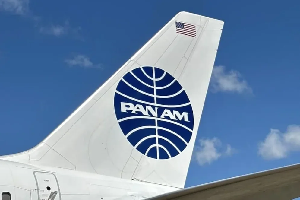 Lego model of a Pan Am airplane with four Lego figures and Pan Am logo on a blue book atop a wicker table next to a lamp and plant.