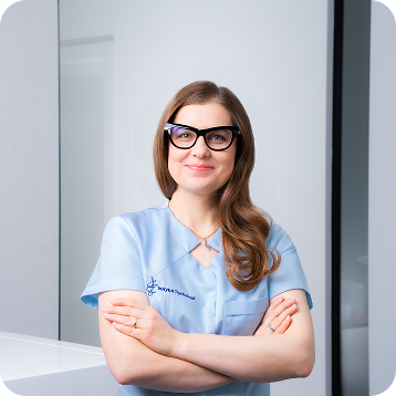 Kobieta w niebieskim uniformie medycznym z założonymi rękami i okularami, stojąca w jasnym, nowoczesnym wnętrzu.