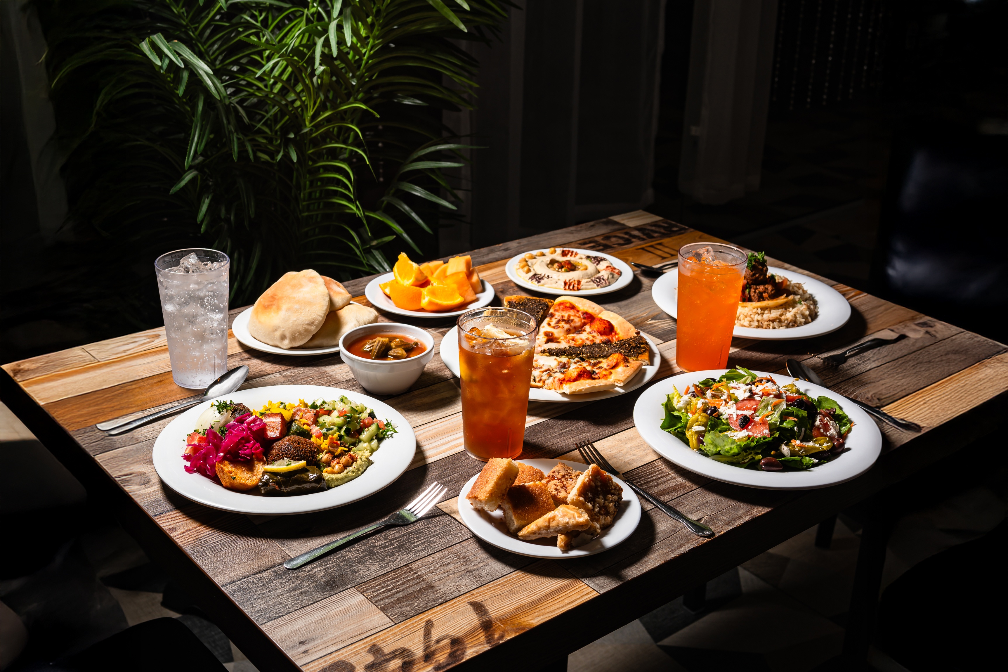 Wooden table set with Mediterranean dishes including salad, pizza, hummus, pita bread, soup, fruit, desserts, and iced drinks.