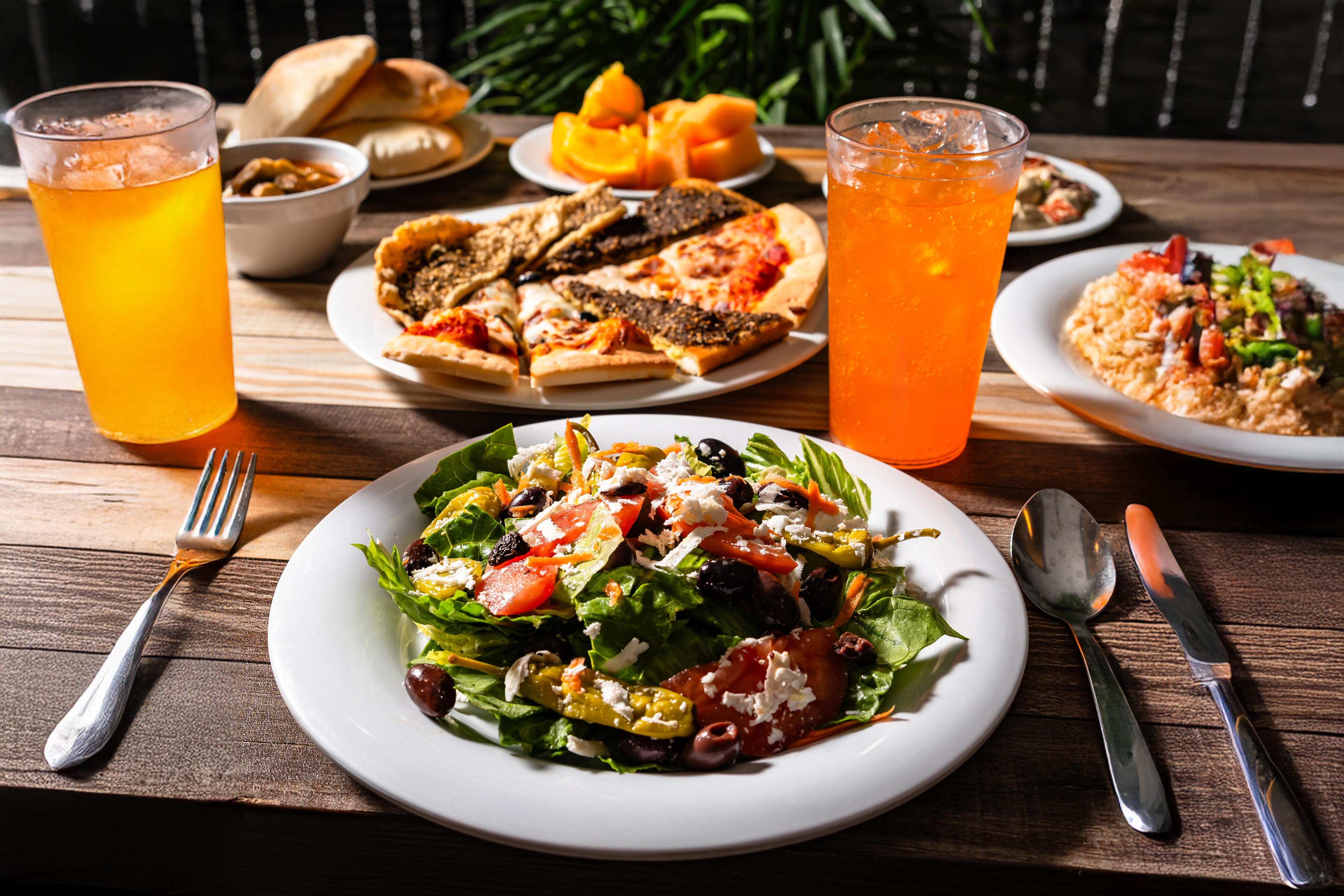 Table setting with a Greek salad, two glasses of orange soda with ice, a plate of pizza slices, bread rolls, fruit pieces, and a dish with rice and vegetables.