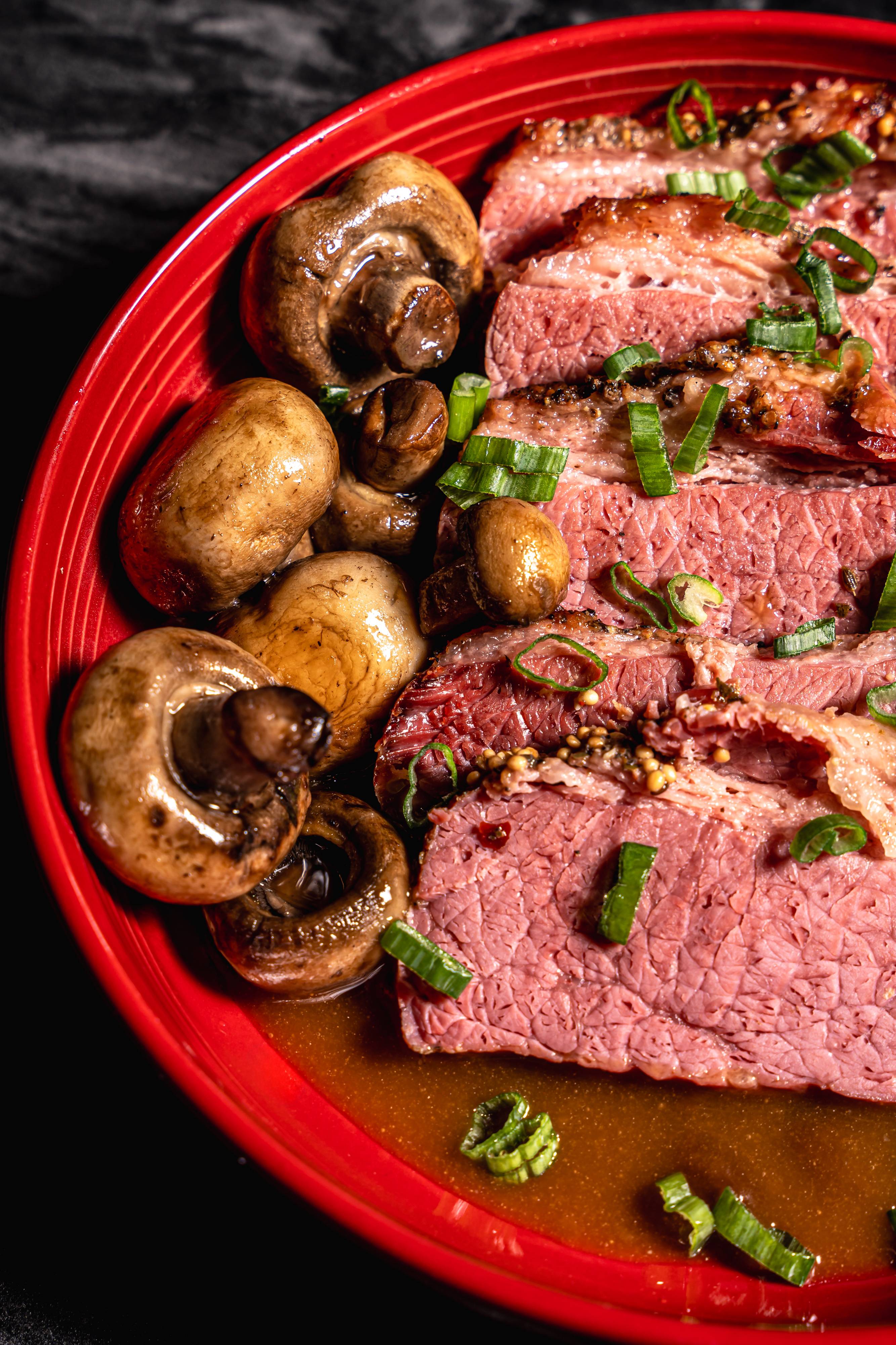 Red plate with sliced cooked brisket topped with chopped green onions and whole cooked mushrooms in broth.