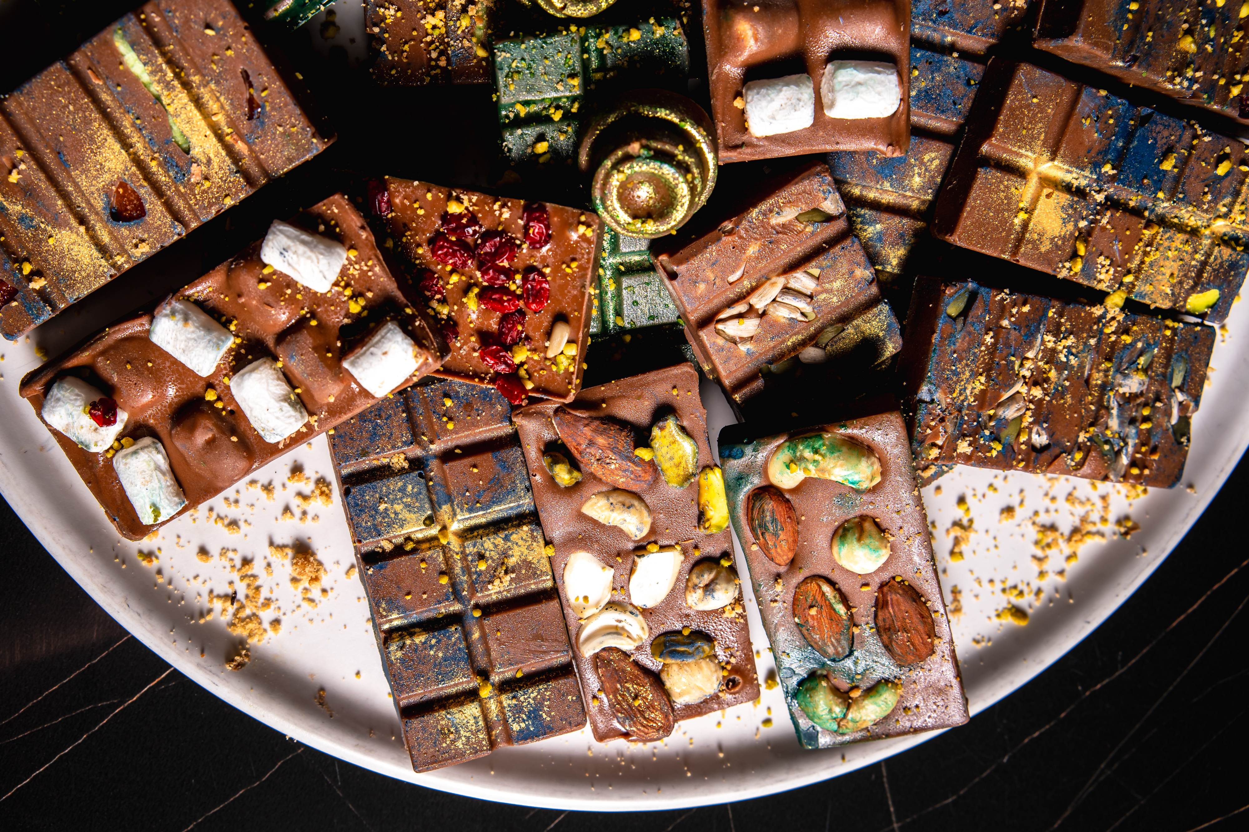 Assorted chocolate bars topped with nuts, seeds, marshmallows, and dried berries on a white plate.