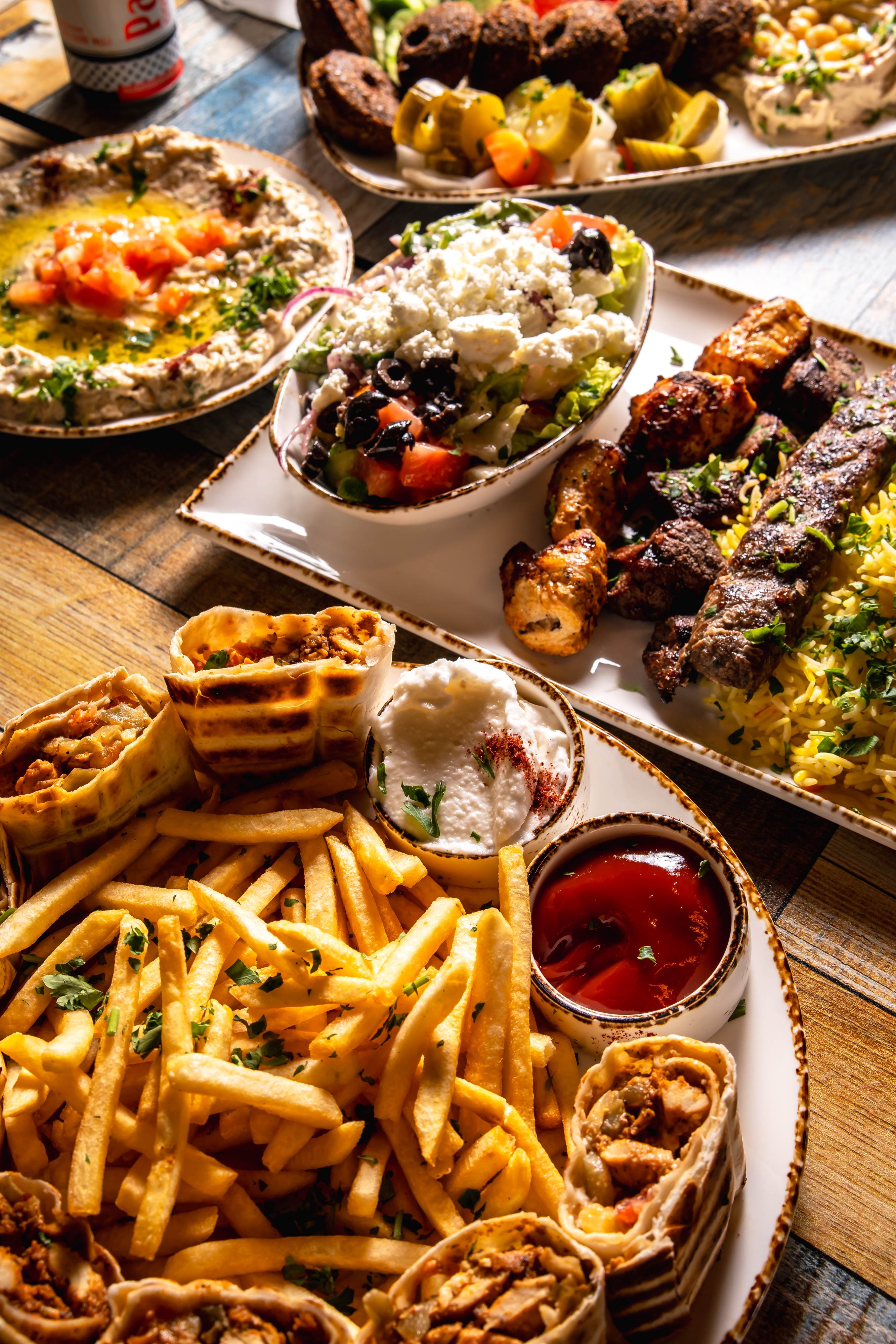 Plates featuring Mediterranean dishes including grilled meat with rice and salad, hummus with diced tomatoes, and wraps with french fries served with sauces.