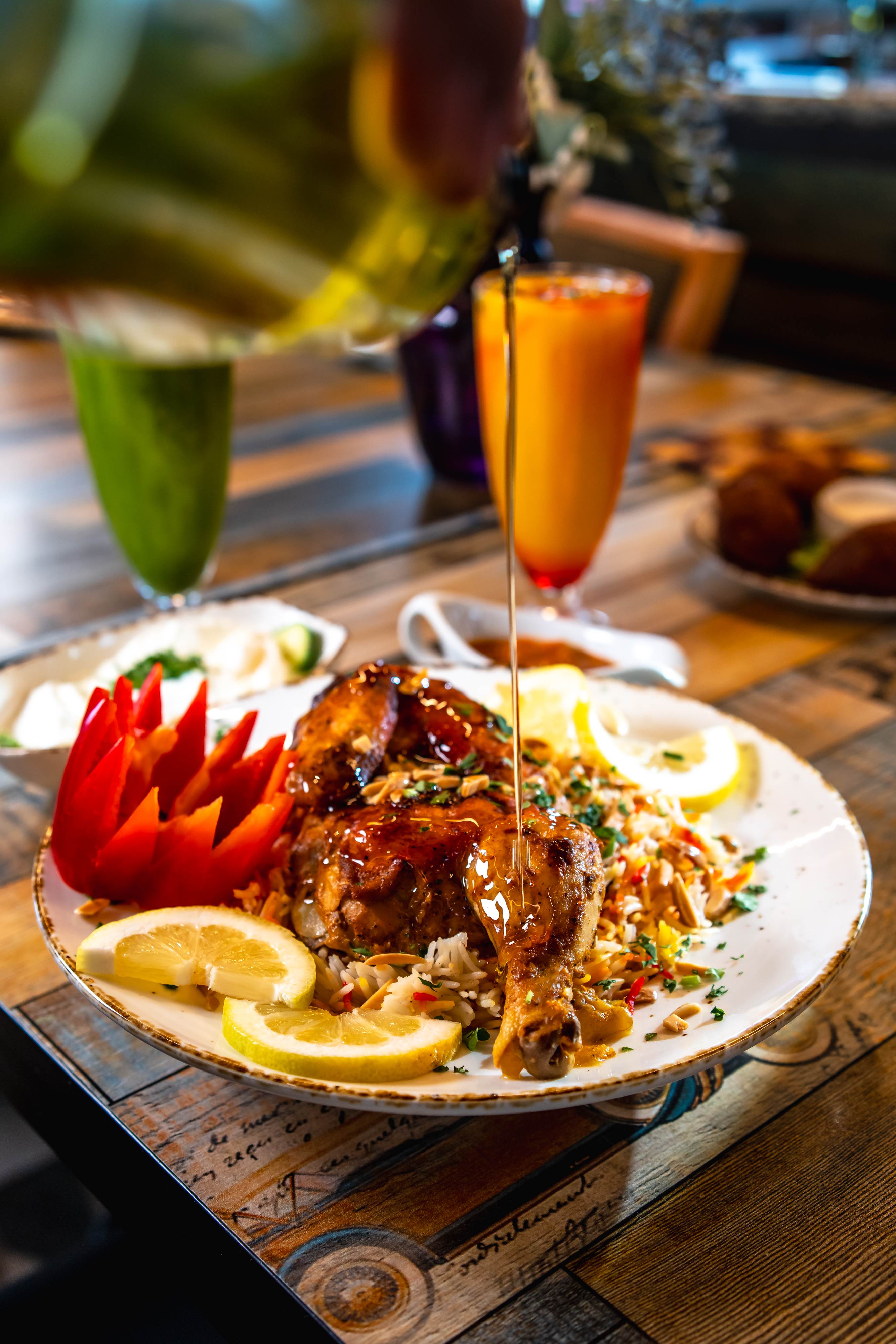 Grilled chicken leg on seasoned rice with almonds, lemon slices, and carved red pepper, drizzled with sauce on a white plate.