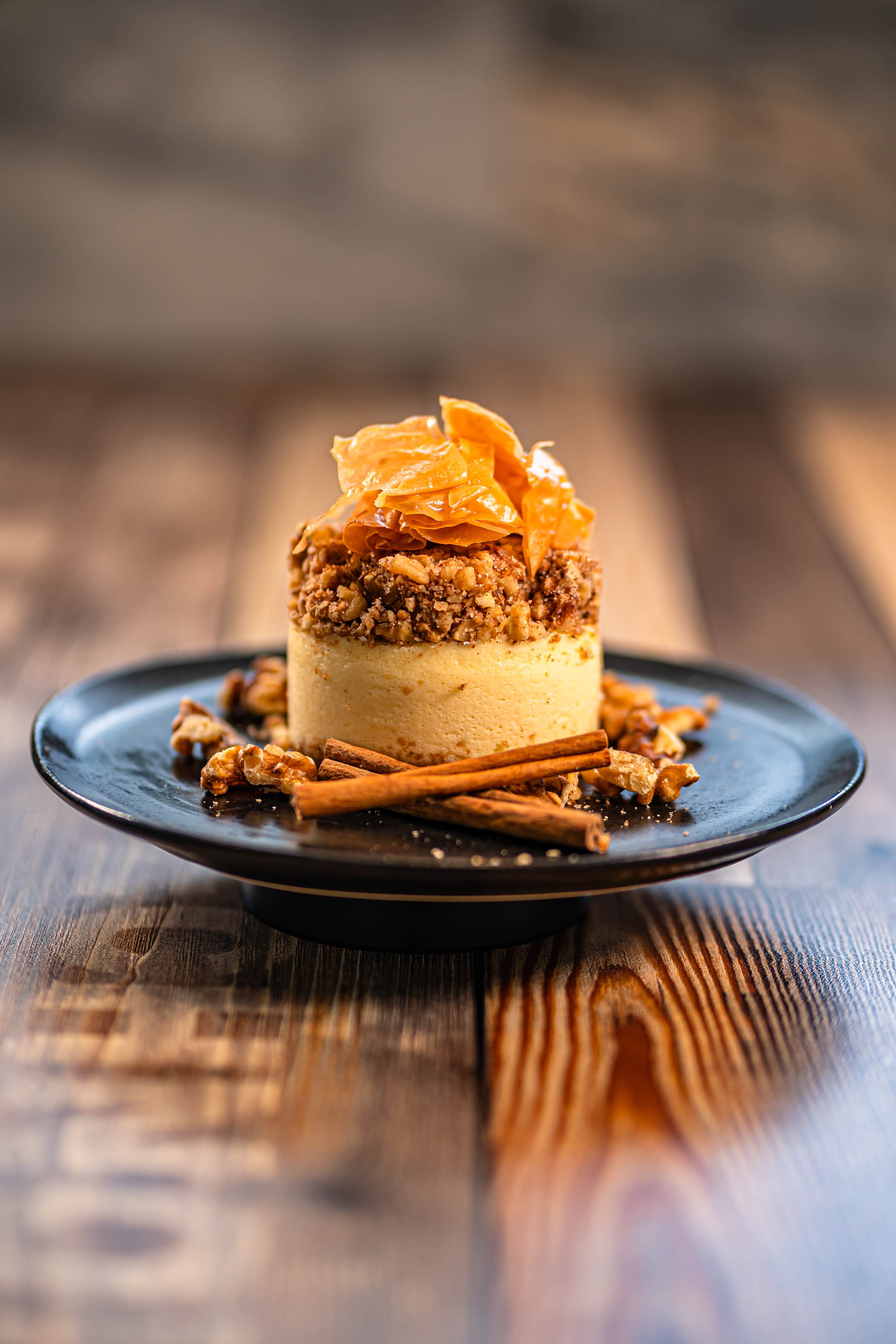 Small round dessert topped with crunchy nuts and thin orange flakes on a black plate with cinnamon sticks and walnuts, on a wooden table.