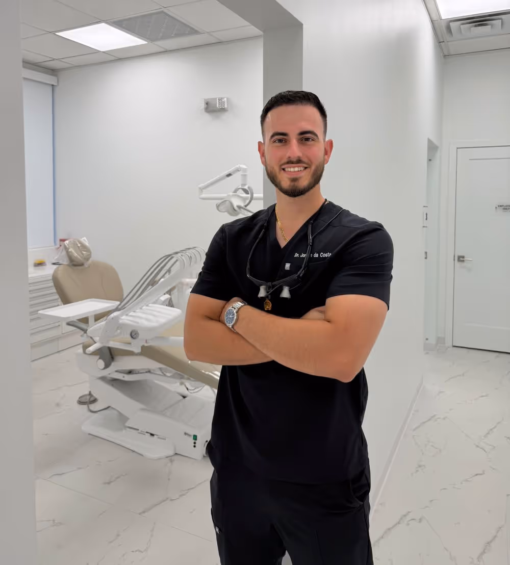 Dentist in black scrubs smiling with arms crossed in a bright, clean dental examination room.