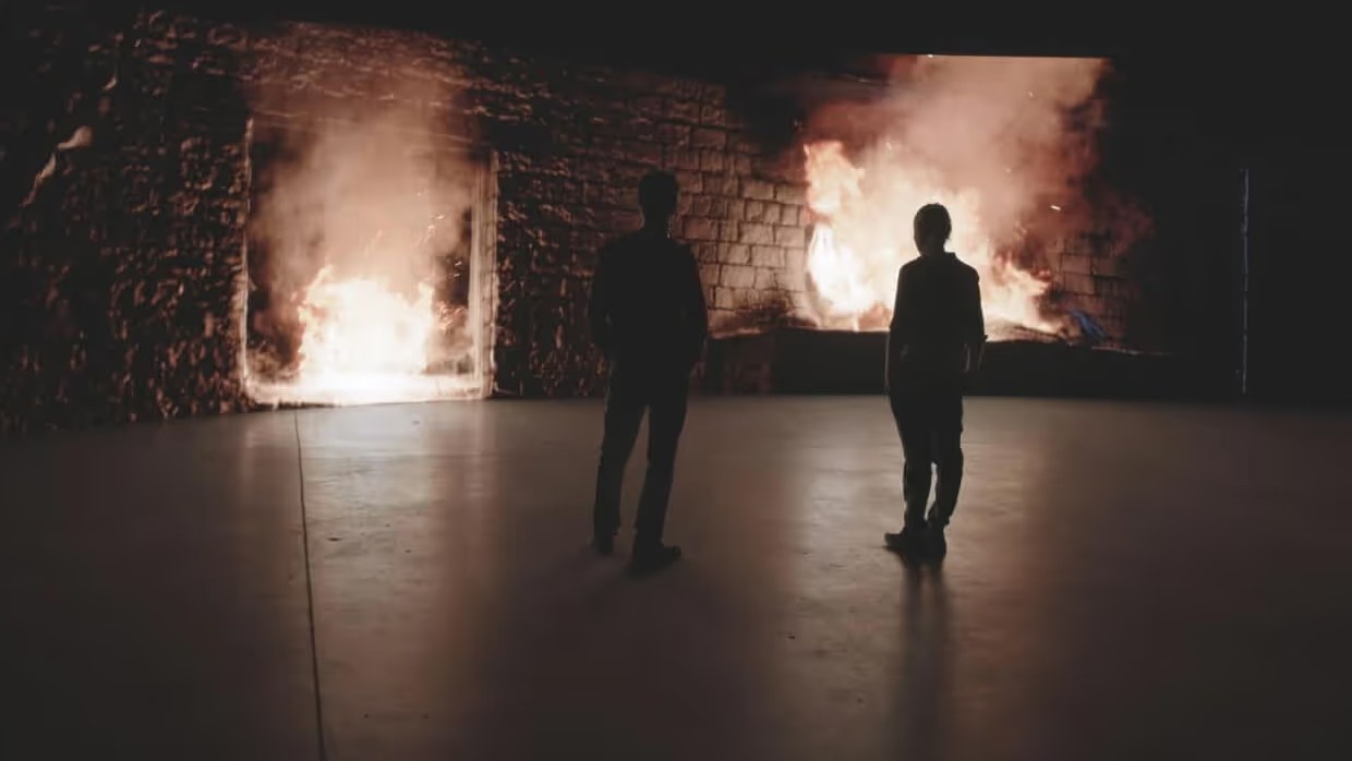 Two silhouettes facing burning ruins inside the Murals installation