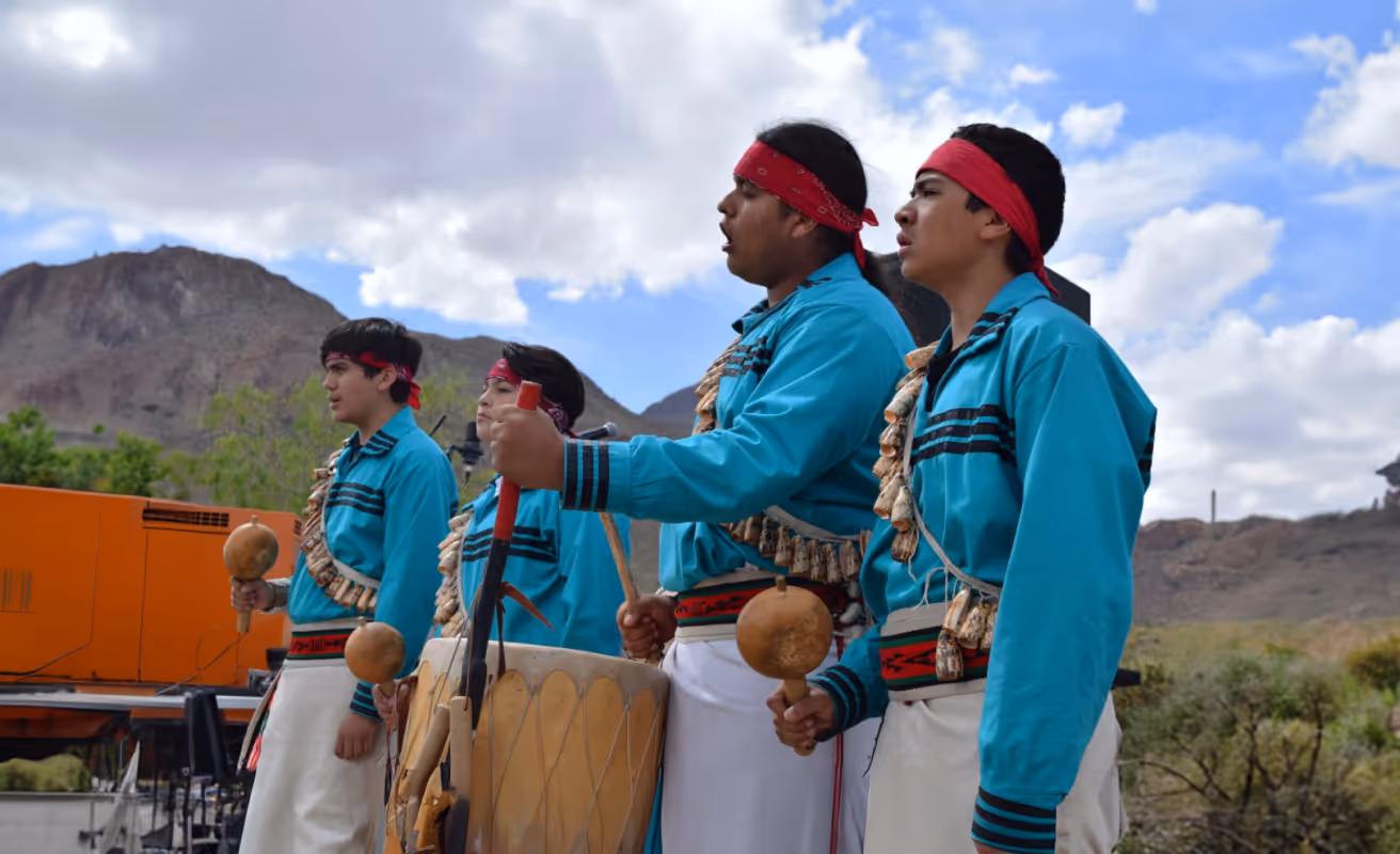 Four men in traditional attire playing drums and rattles outdoors with mountains and cloudy sky in the background.