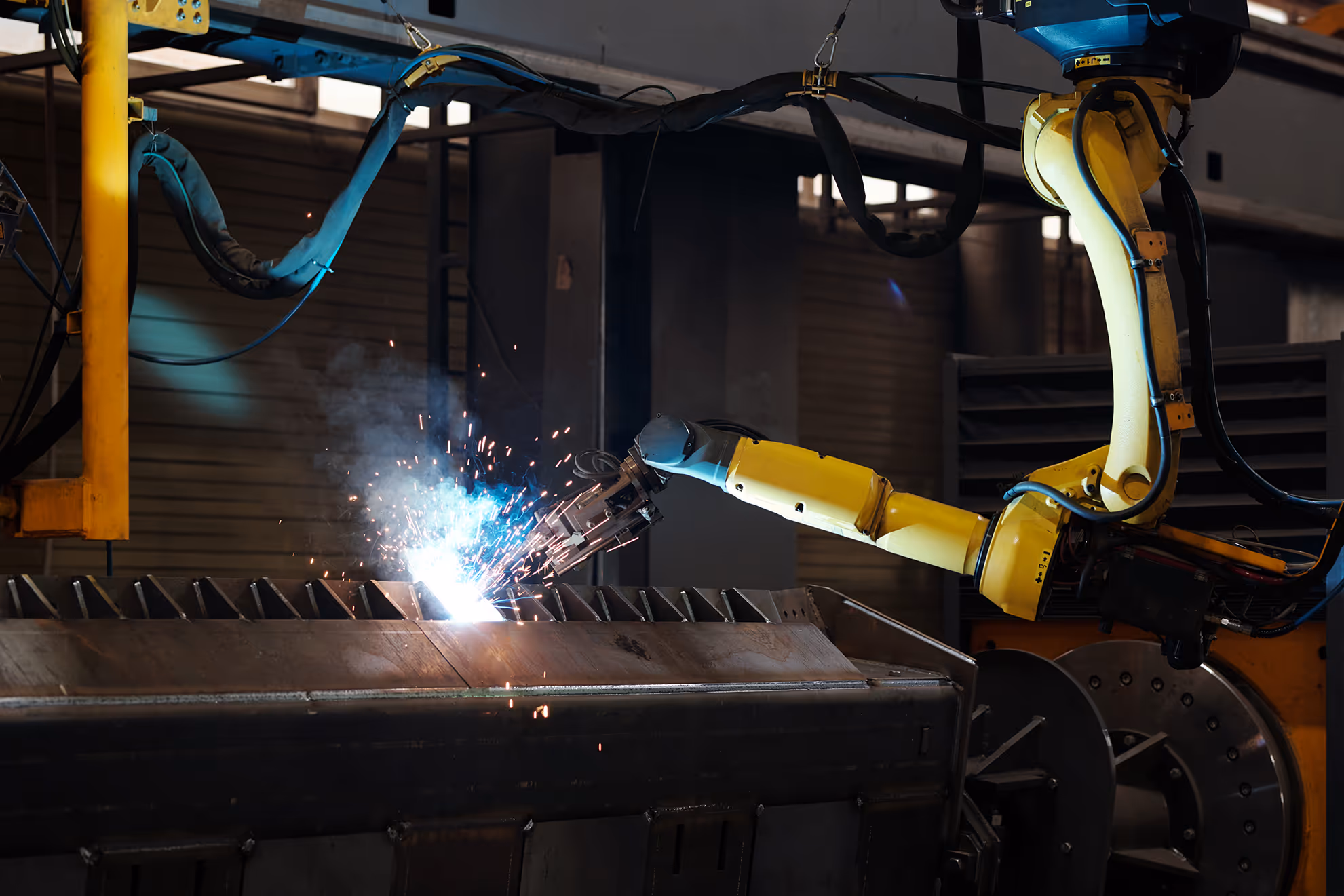Yellow industrial robotic arm welding metal with sparks and bright blue light in a factory setting.
