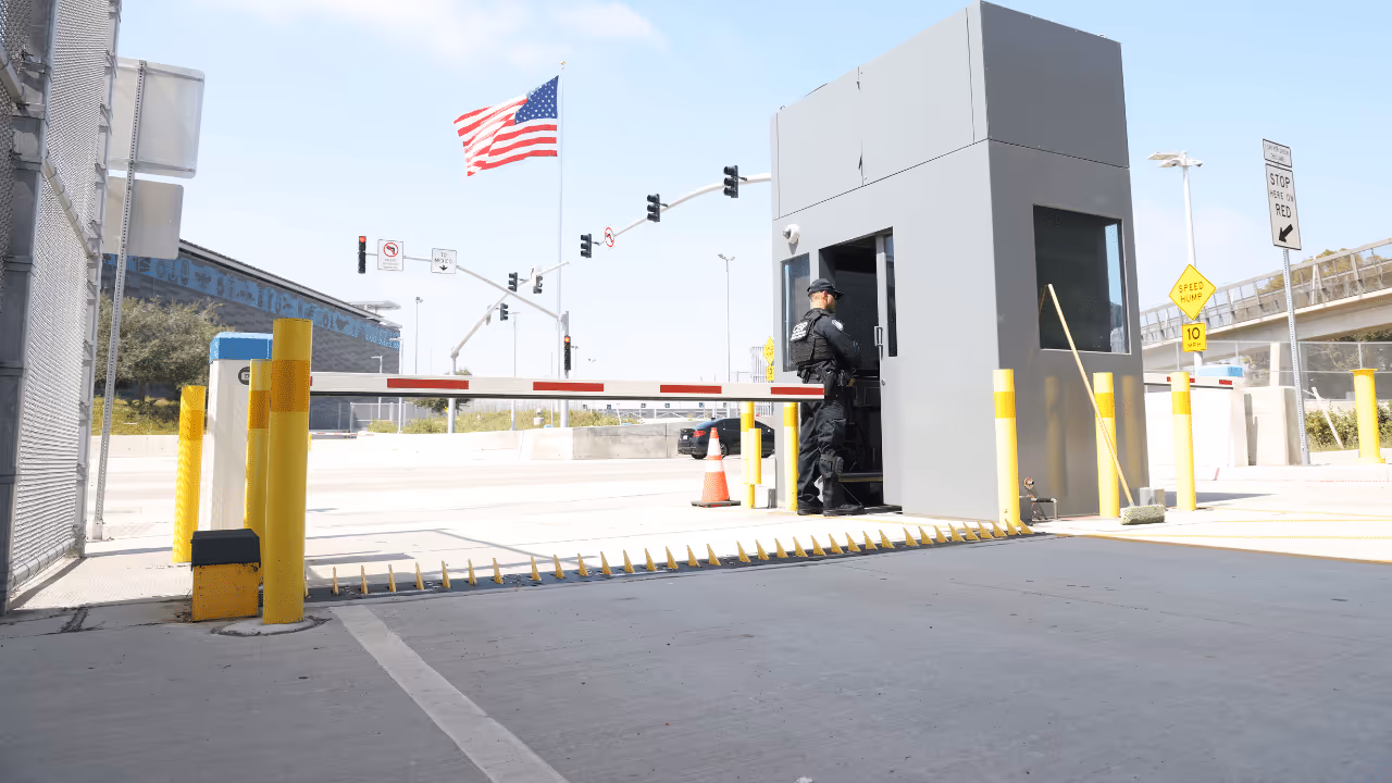 Security guard standing outside a gray checkpoint booth beside a lowered barrier arm and spike strip, with an American flag and traffic lights in the background.