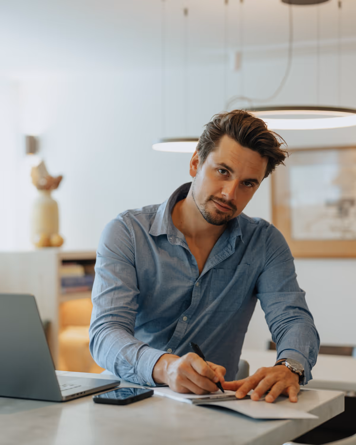 Man in blauw overhemd schrijft aan tafel met laptop en telefoon in een lichte kamer.