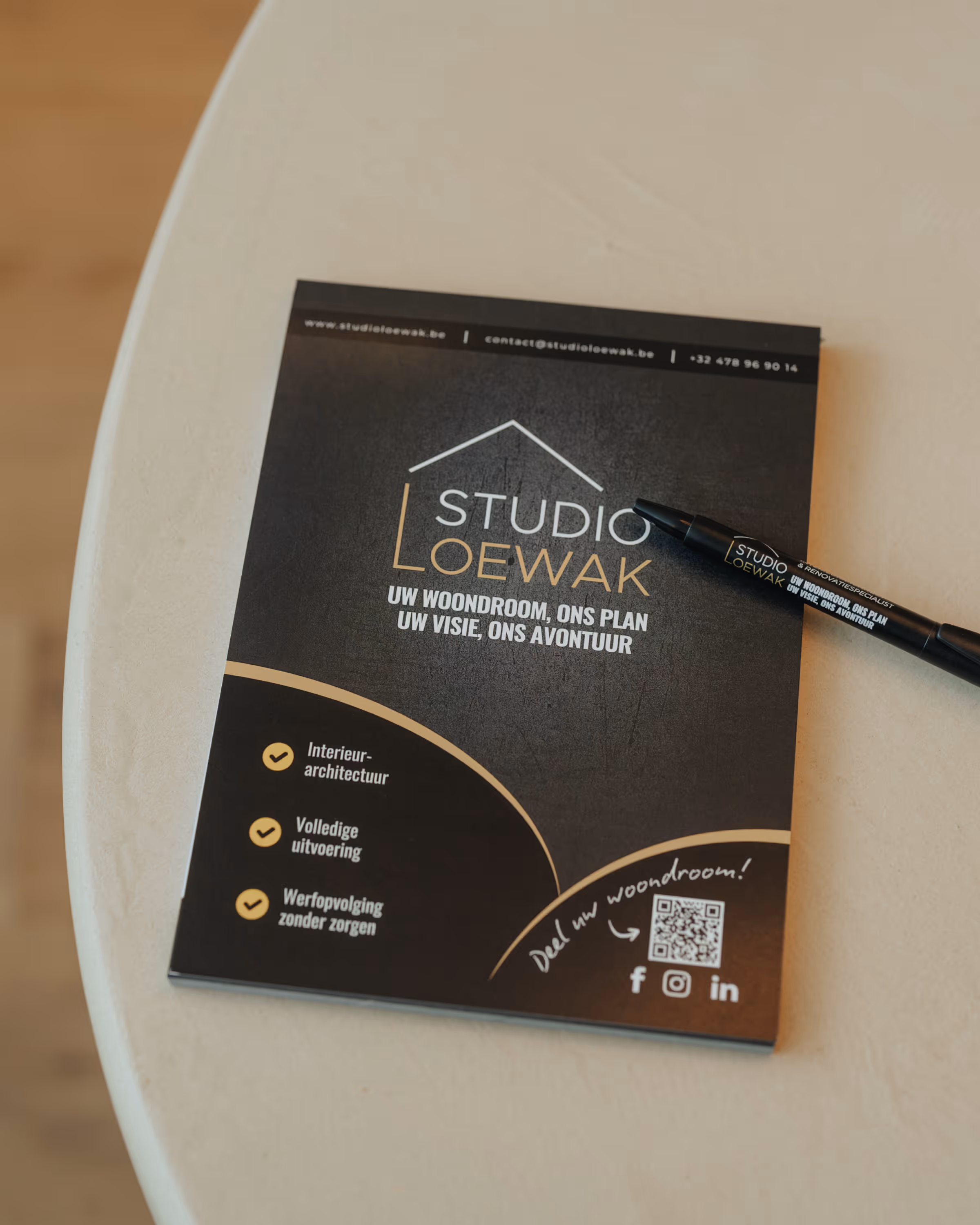 Folder en pen Studio Loewak met slogan, diensten en QR-code op witte ronde tafel.