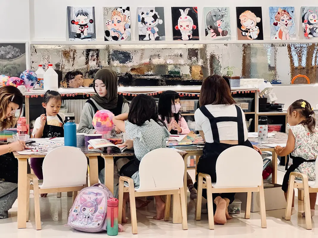 Group of children and two adults seated around a table in an art classroom engaged in creative activities.