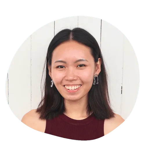 Smiling young woman with straight dark hair wearing silver earrings and a maroon sleeveless top against a white wooden background.