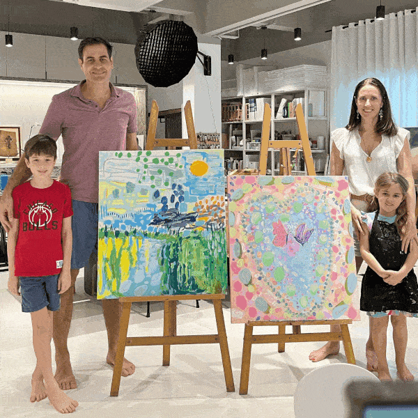 A family of four standing barefoot indoors with two colorful paintings on easels; a man and boy on the left with a landscape painting, a woman and girl on the right with a butterfly-themed painting.