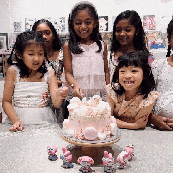 Group of five smiling children gathered around a pink decorated birthday cake with small cartoon character figurines in front on a table.