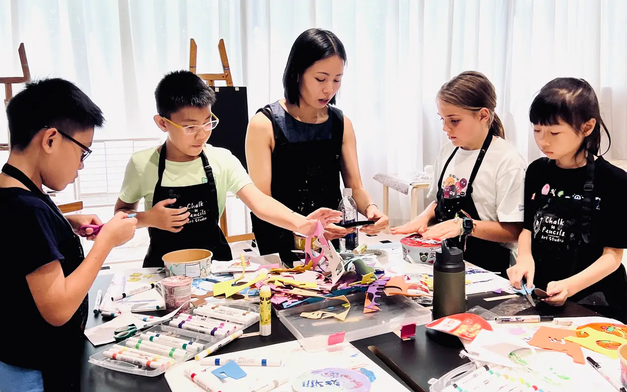 An art teacher guiding four children in a craft activity with scissors, paper, and markers at a table.