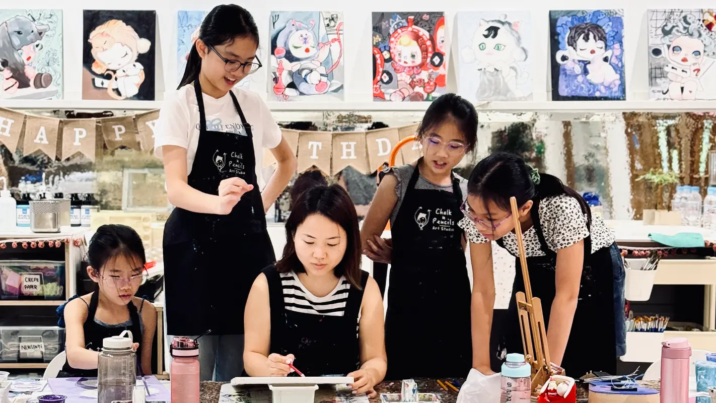 A woman and four girls engaged in a painting class with colorful cartoon-style paintings displayed on the wall behind them.