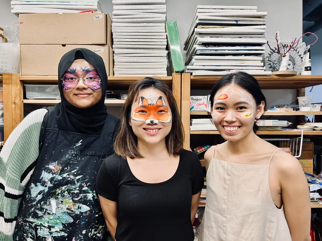 Three women smiling with colorful face paint designs; one wears a black hijab and apron with paint splatters, another has a fox face paint, and the third has citrus fruit designs on her face.