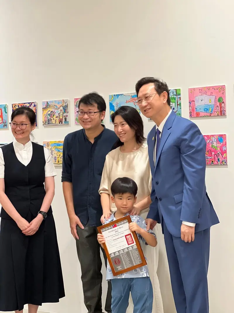 A young boy holding a certificate, standing with three adults smiling in front of a wall decorated with colorful children's drawings.