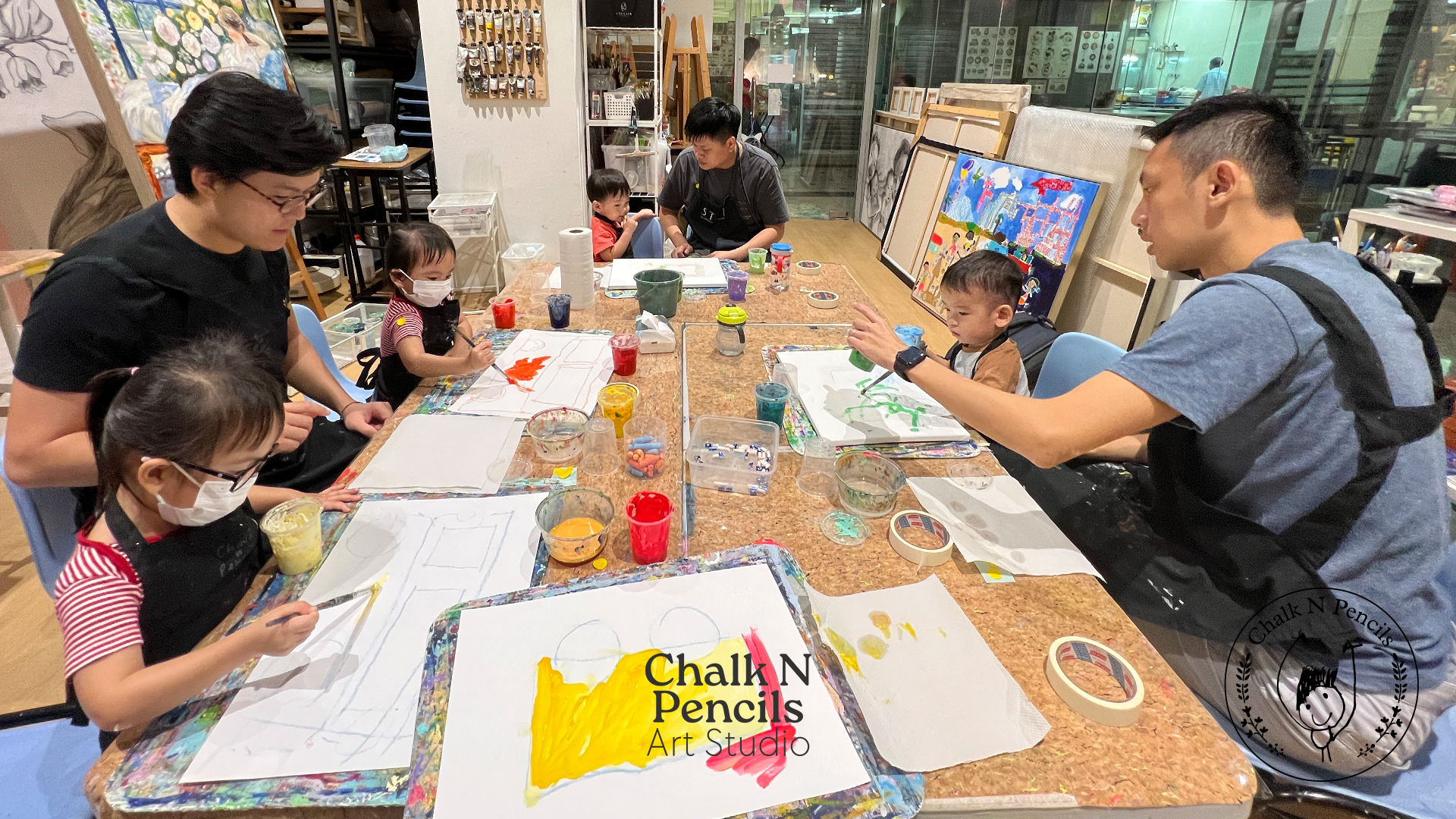Adults and children painting on paper at a table in an art studio with colorful paintings and art supplies around.