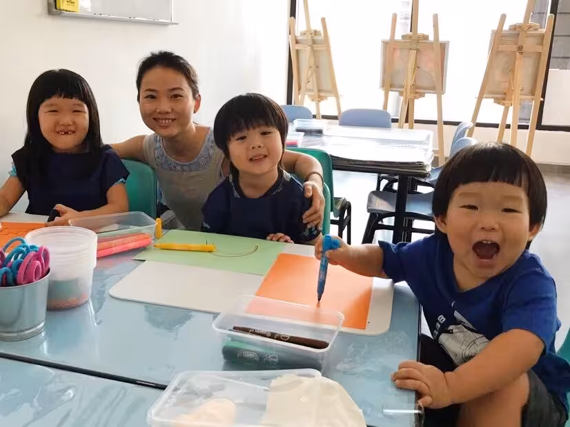 Three young children and a woman at a table with art supplies, with the children smiling and drawing on colored paper.