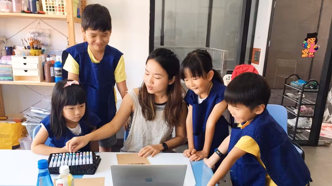 Art teacher guiding four children wearing blue aprons around a table with art supplies and a laptop.