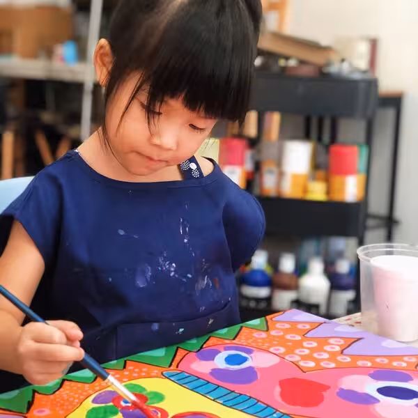 Young girl focused on painting a colorful art piece at a table with paint supplies in the background.