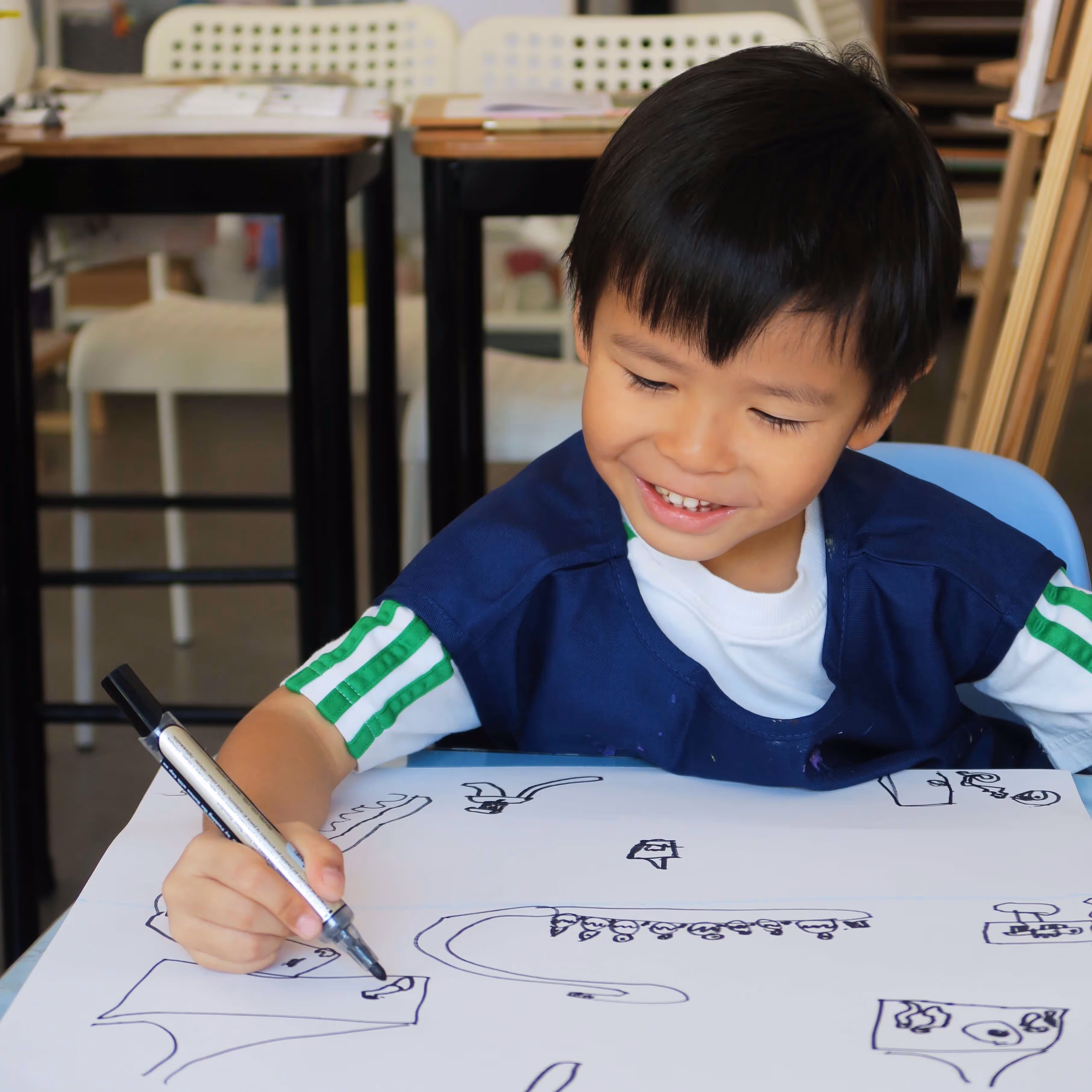 Smiling young boy drawing black marker sketches on white paper at a classroom table.